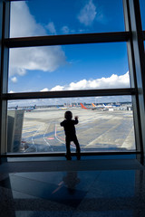 Fototapeta na wymiar A small child looks out of a large window at the airport's tarmac while waiting for his plane