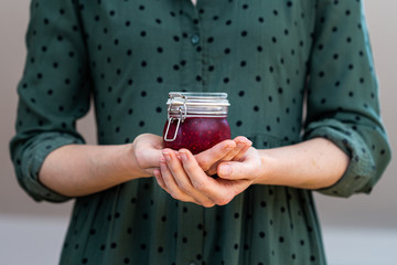 Female hands holding a home made vegan raw raspberry jam in a glass jar with a olive green dress. Healthy raw jam in a cup. 
