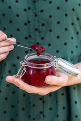 Female hands holding a home made vegan raw raspberry jam in a glass jar with a olive green dress. Healthy raw jam in a cup and silver spoon.