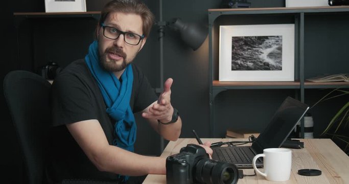 Competent mature photographer in blue scarf and eyeglasses providing online training about retouching images. Bearded man sitting in studio and working on distance.