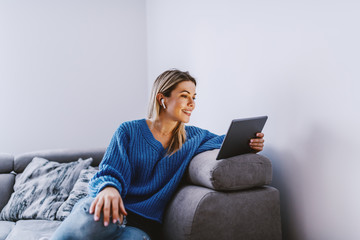 Beautiful caucasian blond woman dressed in blue sweater sitting on sofa in living room and having...