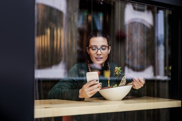 Pensive young girl sitting in restaurant, having salad for lunch and using smart phone for replying...
