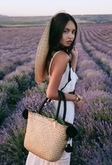 beautiful woman with dark hair in elegant dress and big straw hat posing among flowering lavender...