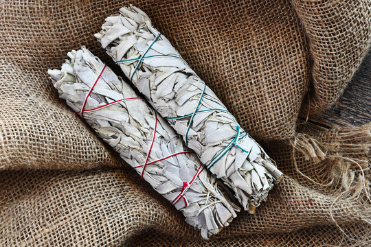 A close up image of two white sage smudge sticks on beige burlap fabric.  