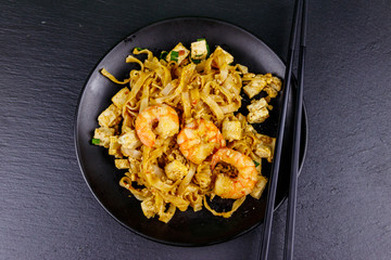Stir-fried rice noodles Pad Thai with prawns and tofu in a plate on black slate background. Top view. Thai food