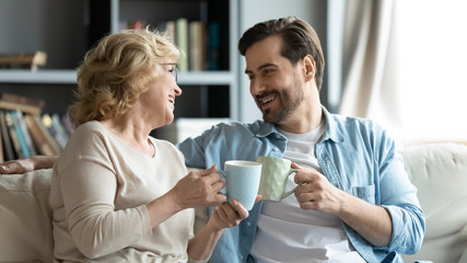 Adult son visit older mother, parent feels happy miss for grown up child enjoy pleasant warm friendly talk at informal atmosphere drinking tea seated on couch in cozy living room, native home concept