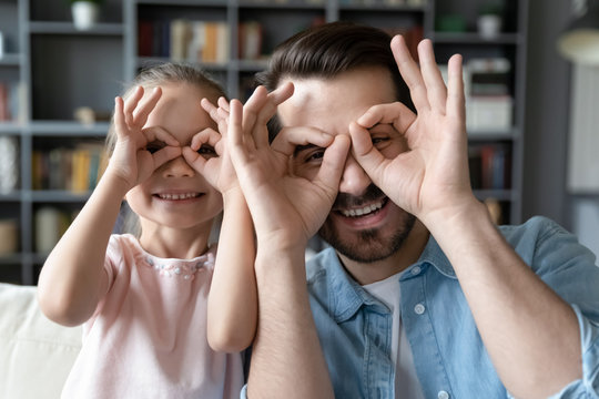 Small daughter and father have fun at home, do funny faces making with fingers eyewear shape like glasses looking at camera through binoculars, eyeglasses sunglasses lenses store advertisement concept