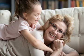 Close up view happy elderly grandma spend time at home with preschooler granddaughter, small kid girl and grandmother lying on couch play laughing enjoy active weekend, love and familial bond concept