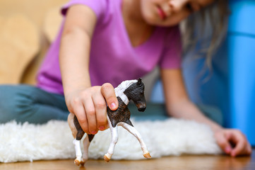 little six year old kid girl playing at home with a children's toy horse sitting on the floor. concept of quarantine self-isolation due to covid-19 corona virus - obrazy, fototapety, plakaty