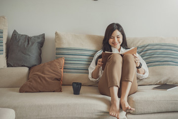 Happy young asian woman reading book sitting on the sofa at home.
