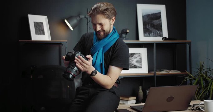 Smiling bearded photographer in stylish outfit leaning on table while reviewing photos on digital camera. Combination of favorite hobby and commercial project.