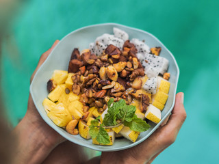 bird view of a vegan fruit porridge bowl with granola, mango, dragon fruit, apple, nuts, cocoa and mint holding in both hands over a turquoise water pool