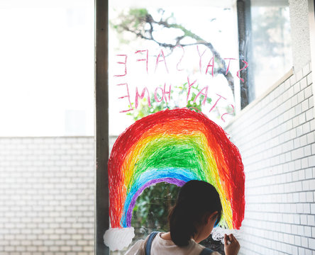 窓に虹と"stay safe, stay home"のメッセージを描く女の子