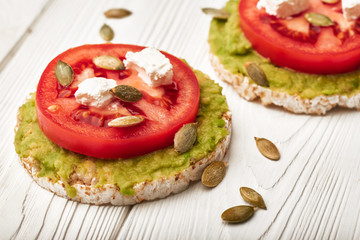 Rice cake with avocado and tomatoes and feta cheese with pumpkin seeds for breakfast on a white wooden background