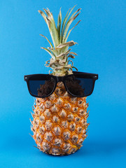 Front view of a ripe orange pineapple with green leaves and black sunglasses on it, on a blue...