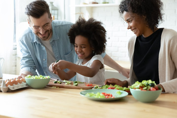Overjoyed international young family with little daughter have fun preparing healthy food salad at home together, happy multiracial parents teach small girl child cooking in kitchen on weekend
