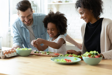 Overjoyed international young family with little daughter have fun preparing healthy food salad at...