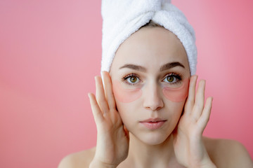 Studio shot of satisfied caucasian freckled woman wearing white towel on head, with collagen patches under eyes, standing naked against pink background. Skin care, cosmetic product concept