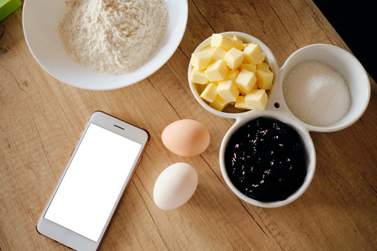 Ingredients For Cake On Table Ready For Cooking While Following A Recipe On Smart Phone With White Screen. Butter, Gem, Eggs, Floor