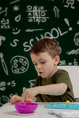 Cute little boy moulds from plasticine on table with a blackboard on a background. Ready for school. Home schooling. Back to school