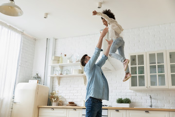 Young Caucasian dad play throw in air excited African American little daughter, happy father have fun engaged in funny activity with biracial small girl child at home kitchen on weekend together