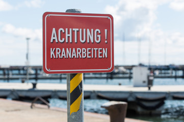 a red sign "Attention! Crane work" (German "Achtung! Kranarbeiten") in front of crane constructions on a quayside