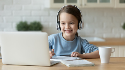 Smiling little Caucasian girl in headphones handwrite study online using laptop at home, cute happy small child in earphones take Internet web lesson or class on computer, homeschooling concept - Powered by Adobe