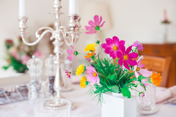 a table set for a dinner in a light room, covered with a white tablecloth, decorated with silver candlestick and a bouquet of colorful garden flowers 