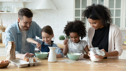 Loving happy diverse parents with little multiracial daughters baking in modern kitchen, smiling multiethnic young family with small girls children cook together prepare pancakes or biscuits at home