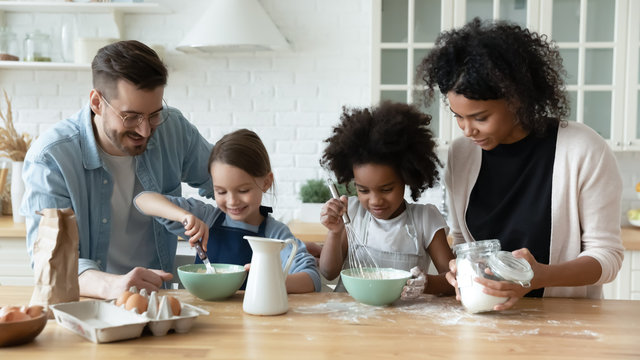 Loving happy diverse parents with little multiracial daughters baking in modern kitchen, smiling multiethnic young family with small girls children cook together prepare pancakes or biscuits at home - Powered by Adobe