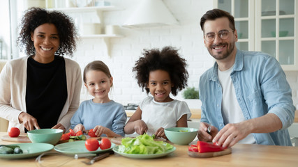 Portrait of happy young multiracial family with little children cook together in modern renovated kitchen, smiling multiethnic parents have fun preparing healthy food breakfast with small daughters