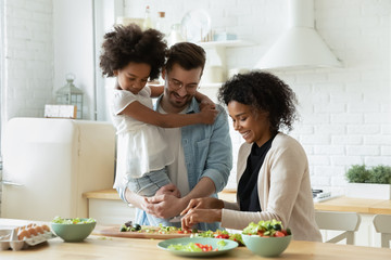 Happy young diverse parents have fun teach little biracial daughter cooking, overjoyed multiracial family with small girl child preparing food making healthy salad for breakfast in kitchen together - Powered by Adobe
