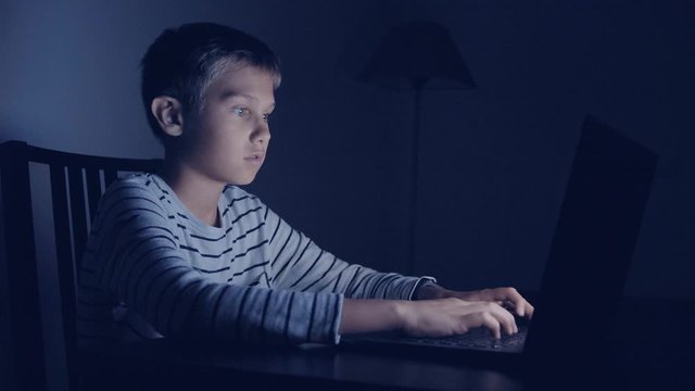 Boy using laptop computer at home at night. Young boy playing computer video games, surf on net