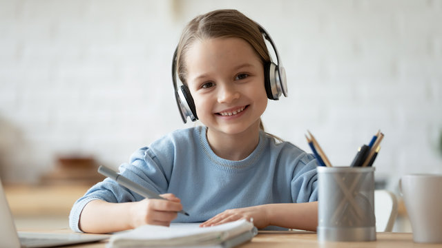 Portrait of smiling little girl wearing wireless headphones drawing at home, happy small preschooler child in earphones have fun painting picture on weekend, studying online on quarantine