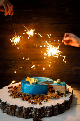 Blue Birthday cake with colorful decor and firework sparkles in hands over a wood background.