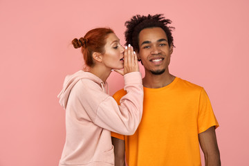 Young ginger woman whispers secret to dark-skinned boyfriend who has cheerful expression, gossip together, wear casual clothes, stands against pink wall. Diverse couple indoor. Secret, gossip concept