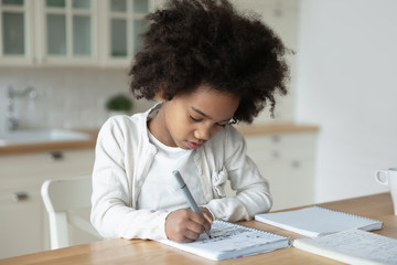 Small African American girl sit at desk in kitchen handwrite in notebook having distant class, smart little biracial kid prepare homework study alone on quarantine at home, homeschooling concept