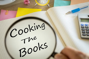 Magnifying glass writing 'Cooking the books' in a notebook.  Creative accounting concept.  Calculator, office accessories, clock