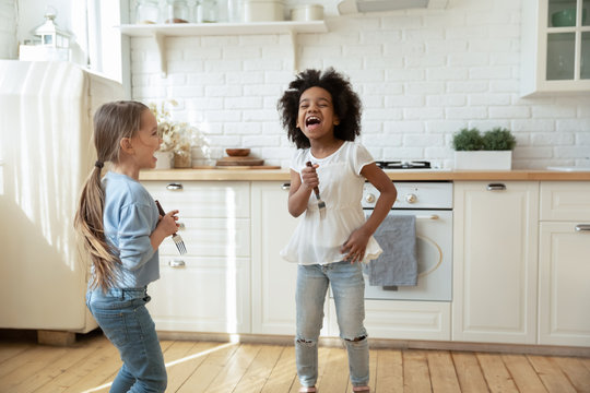 Overjoyed little multiracial sisters have fun play singing in kitchen appliances together, happy small multiethnic girls siblings engaged in funny activity at home enjoy leisure weekend or quarantine