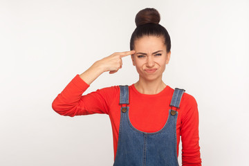 Crazy cuckoo mind. Portrait of girl with hair bun in denim overalls showing stupid gesture and looking with displeased annoyed face, accusing insane idea. studio shot isolated on white background