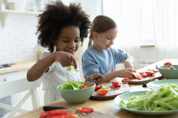 Excited little multiracial girls sisters have fun chopping vegetables making healthy salad at home, happy small multiethnic siblings children cooking in kitchen together, preparing food for parents