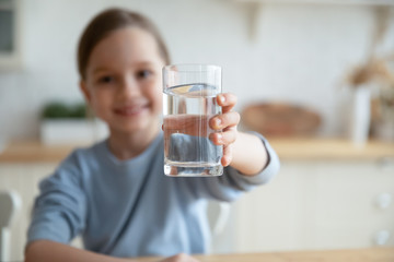 Close up of happy little Caucasian girl offer crystal still mineral water for body refreshment,...