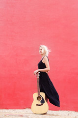 Vertical photo of a young beautiful blond woman in a black dress standing leaning on a guitar against a red wall background. Empty space on top for text