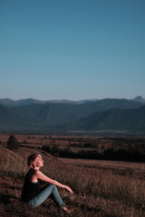 Vertical shot - a young woman sits on a hill at sunset and looks peacefully and relaxed at the mountains. Empty space on top for text