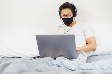 Man with protective mask and headphone working on laptop in bed at home covid-19 outbreak.