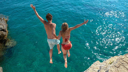 Joyful tourist couple decides to jump off a rocky cliff and dive into sea. - obrazy, fototapety, plakaty
