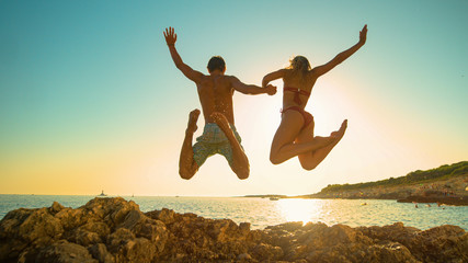 LOW ANGLE: Cheerful young couple holds hands while diving into ocean at sunset - obrazy, fototapety, plakaty