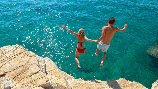 Carefree young tourists holds hands while jumping into the refreshing blue sea.