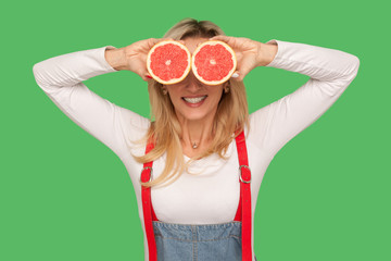 Fresh fruits, natural organic food. Portrait of happy stylish woman covering eyes with sliced juicy grapefruits and smiling, vitamins for healthy diet. indoor studio shot isolated on green background