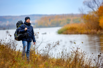 A young man with rucksack standing next to the river. Good warm autumn weather for hiking.